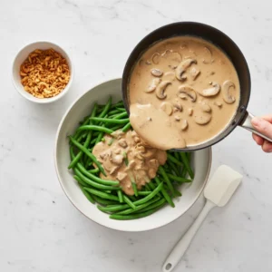 A hand pouring a rich, creamy mushroom sauce from a skillet over a large bowl of blanched fresh green beans to assemble a homemade green bean casserole.