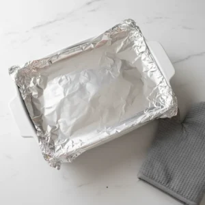 A 9x13 white baking dish filled with a homemade stuffing recipe, covered tightly with aluminum foil before its first bake.