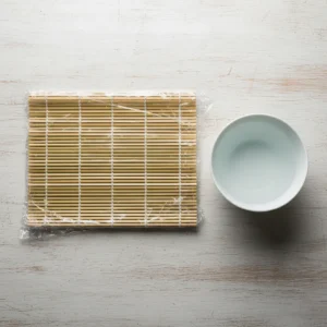 A top-down view of a sushi-making station, showing a bamboo rolling mat wrapped in plastic and a small bowl of water for hands.