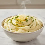 A steaming, large bowl of smooth, creamy mashed potatoes garnished with melted butter, chopped chives, and fresh herbs, ready to be served.