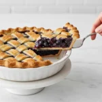 A hand serving a slice of a homemade blueberry pie with a golden lattice crust and thick, juicy filling from a white dish on a pedestal stand, representing the final Blueberry Pie Recipe.