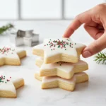 A hand stacking beautifully iced star-shaped Christmas Cookies with red and green sprinkles on a marble counter, with a metal cookie cutter in the background.