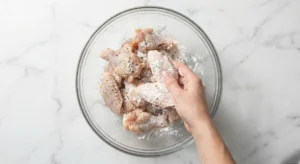 A hand tossing raw chicken wings in a glass bowl to coat them evenly in the baking powder mixture, the key hack for extra crispy buffalo wings oven style.