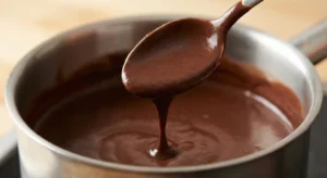 A close-up of a wooden spoon lifted from a pot, coated in a thick, velvety, dark brown chocolate mixture that drips slowly, showing a pudding-like texture.