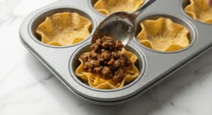 A silver spoon placing a portion of seasoned, browned taco meat into a tortilla cup sitting in a grey muffin tin, filling the mini loaded nacho cups.