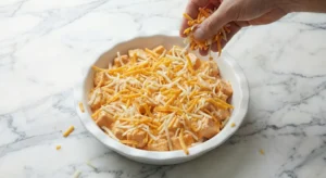 A hand generously sprinkling shredded cheddar and mozzarella cheese over the assembled buffalo chicken dip recipe mixture in a white pie plate, preparing the final crust.