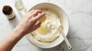 A close-up shot of a hand pouring liquid ingredients from a small glass bowl into a stand mixer bowl filled with the creamed butter and sugar mixture, with the paddle attachment stationary.