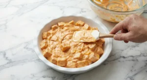 A hand using a spatula to spread the assembled creamy buffalo chicken dip mixture, complete with chicken chunks, evenly into a white pie plate, preparing for baking.