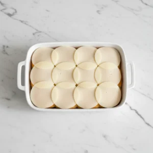A white baking dish containing the bottom slab of toasted rolls completely covered with a layer of overlapping sliced white cheese, creating a barrier for the pizza sauce