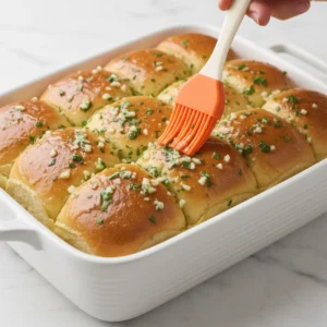 A hand using an orange silicone brush to generously coat the tops of the golden brown rolls with a herb garlic butter mixture, completing the pull apart pizza sliders.