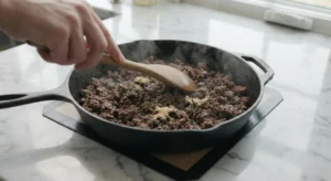A close-up of a hand using a wooden spoon to stir and break apart browning ground beef in a black cast iron skillet on a white marble countertop.