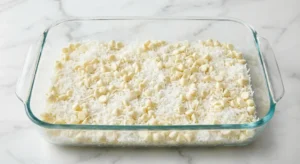A clear 9x13 inch glass baking dish resting on a white marble countertop, with its bottom evenly covered in a thick layer of shredded white coconut and white chocolate chips.