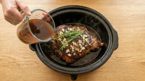 A hand pouring dark beef broth from a glass measuring cup around a seared leg of lamb in a slow cooker.