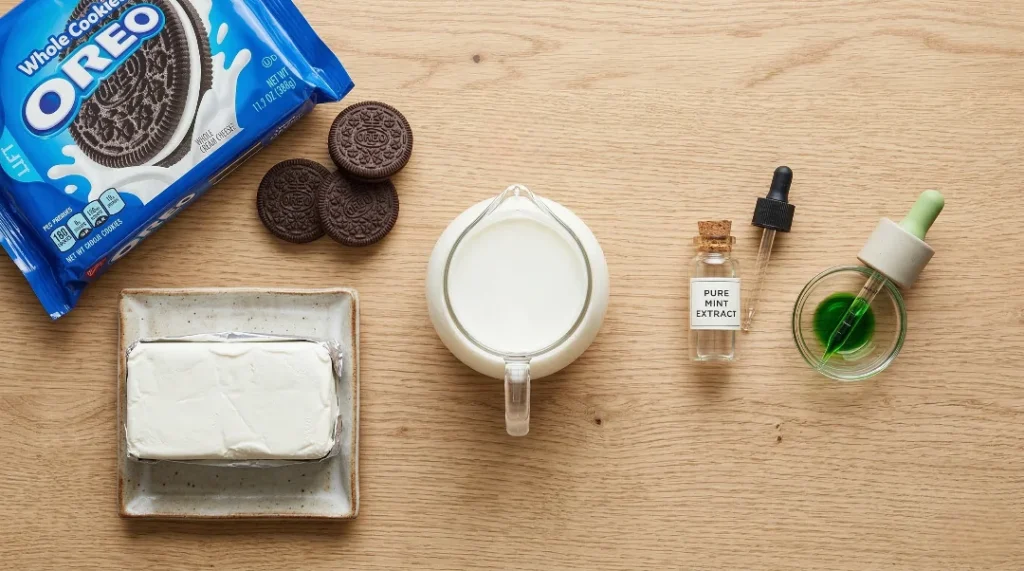 A top-down view of Oreo cookies, a block of cream cheese, a jug of milk, mint extract, and green food coloring on a wooden table.