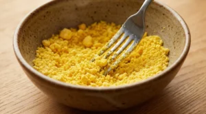 A silver fork mashing bright yellow egg yolks into fine crumbs inside a rustic ceramic bowl on a wooden table.