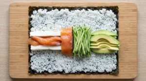 Philadelphia roll recipe assembly showing a row of cream cheese, smoked salmon, cucumber, and avocado arranged horizontally on sushi rice.