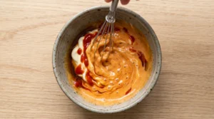 Shrimp tempura roll sauce being made by whisking together creamy Kewpie mayo, sriracha, and sesame oil in a small ceramic bowl.