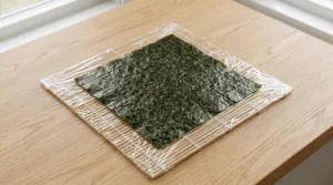 Shrimp tempura roll recipe preparation showing a bamboo sushi mat covered in clear plastic wrap with a dark green nori sheet placed on top.