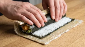 Shrimp tempura roll recipe step-by-step showing hands using a plastic-wrapped bamboo mat to firmly roll nori and rice over shrimp and cucumber fillings.