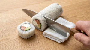 Philadelphia roll recipe step showing a sharp knife slicing an inside-out sushi roll on a wooden board next to a damp white cloth.