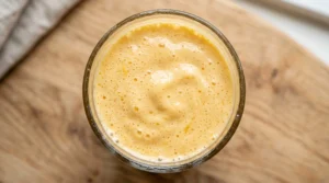 Easy mango lassi recipe top-down view showing thick frothy beverage texture in a glass on a light oak wood table.