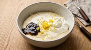 New york cheesecake with sour cream batter step showing sour cream, heavy cream, vanilla bean paste, and lemon zest added to a rustic bowl.