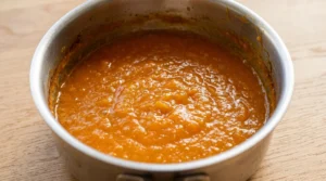 No churn mango ice cream prep showing thick reduced bright orange mango puree in a silver saucepan on a light oak wood table.