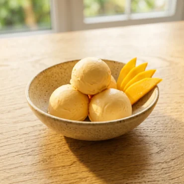 No-Churn Mango Ice Cream recipe cover photo featuring perfectly round scoops in a speckled bowl with fresh mango slices on an oak table.