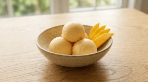 Mango ice cream recipe final presentation showing three perfect scoops in a speckled ceramic bowl with fresh mango slices on an oak table.