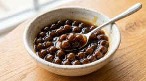 To show the best jasmine bubble tea recipe, glossy tapioca pearls soaked in brown sugar sit in a ceramic bowl on a light oak wood table. Bright natural window light highlights the chewy texture. We bend the strictly zero utensils rule here for a silver spoon to show the perfect soak. Getting the right boil and simmer is the quick and easy way to prep your boba pearls at home. This step is vital for your Viral Food Hacks fans everywhere