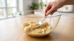 Banana chocolate chip muffins with ripe bananas preparation showing a hand using a metal fork to mash peeled bananas thoroughly inside a large clear glass bowl on a wooden kitchen counter.