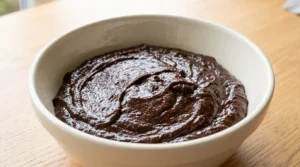 Homemade brownie cookies recipe showing a bowl of rich, thick chocolate batter resting and firming up on a light oak wooden surface.